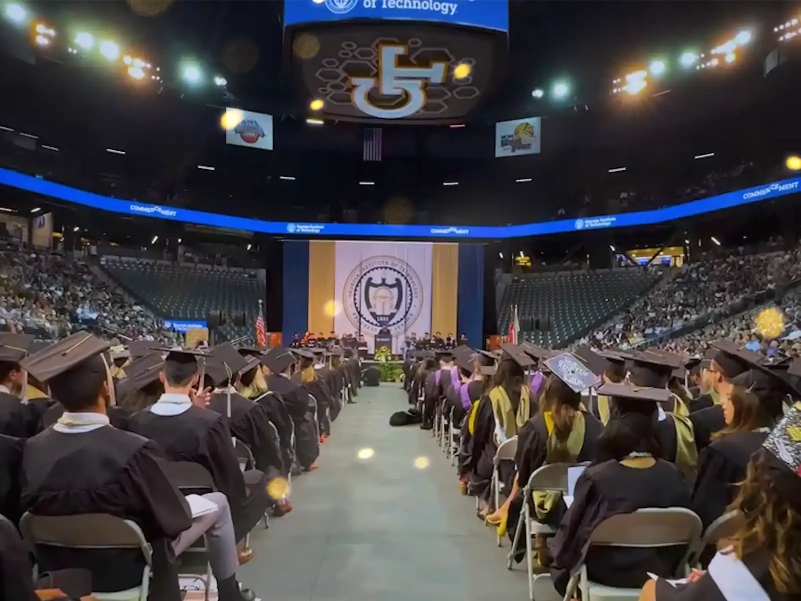 Graduates in caps and gowns sit in an arena facing a stage with the Georgia Tech seal during a commencement ceremony.