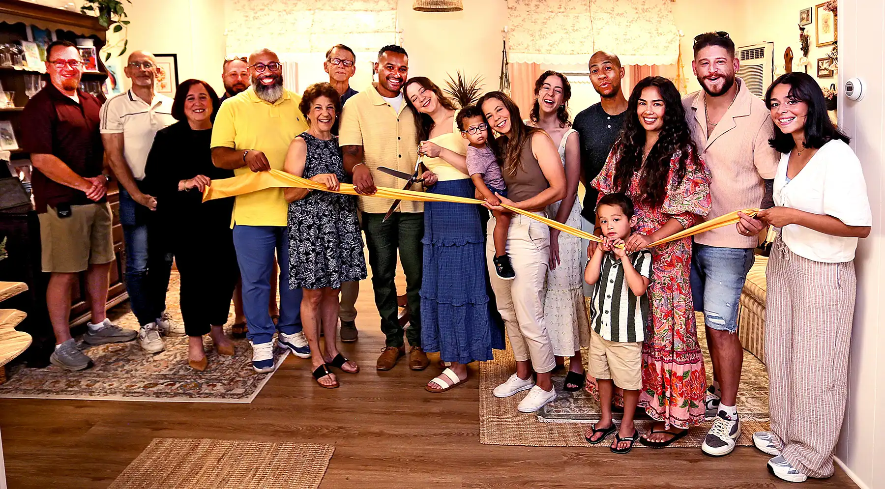A large, joyous group of friends, family, and local officials standing in a row indoors while holding a long, bright yellow grand opening ribbon.