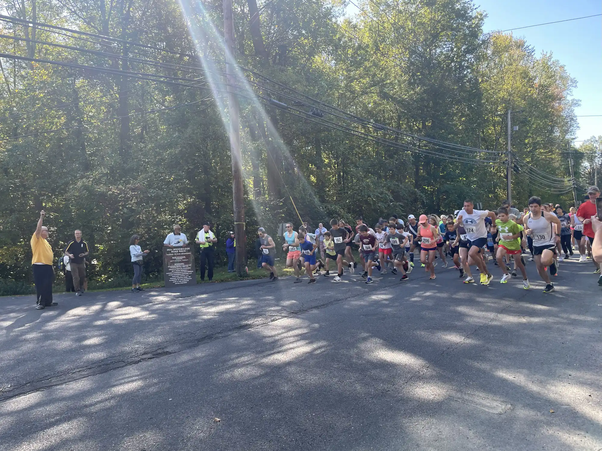 Runners at the starting line of the annual Woodbridge 5K Road Race running along a tree-lined street on a bright, sunny morning outdoors.