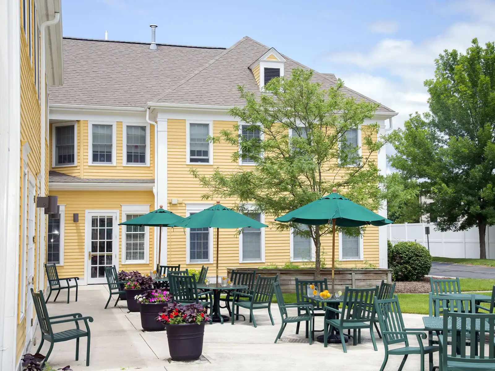 Outdoor patio at Coachman Square in Woodbridge with green tables, patio umbrellas, potted flowers, and chairs beside a yellow clapboard building.