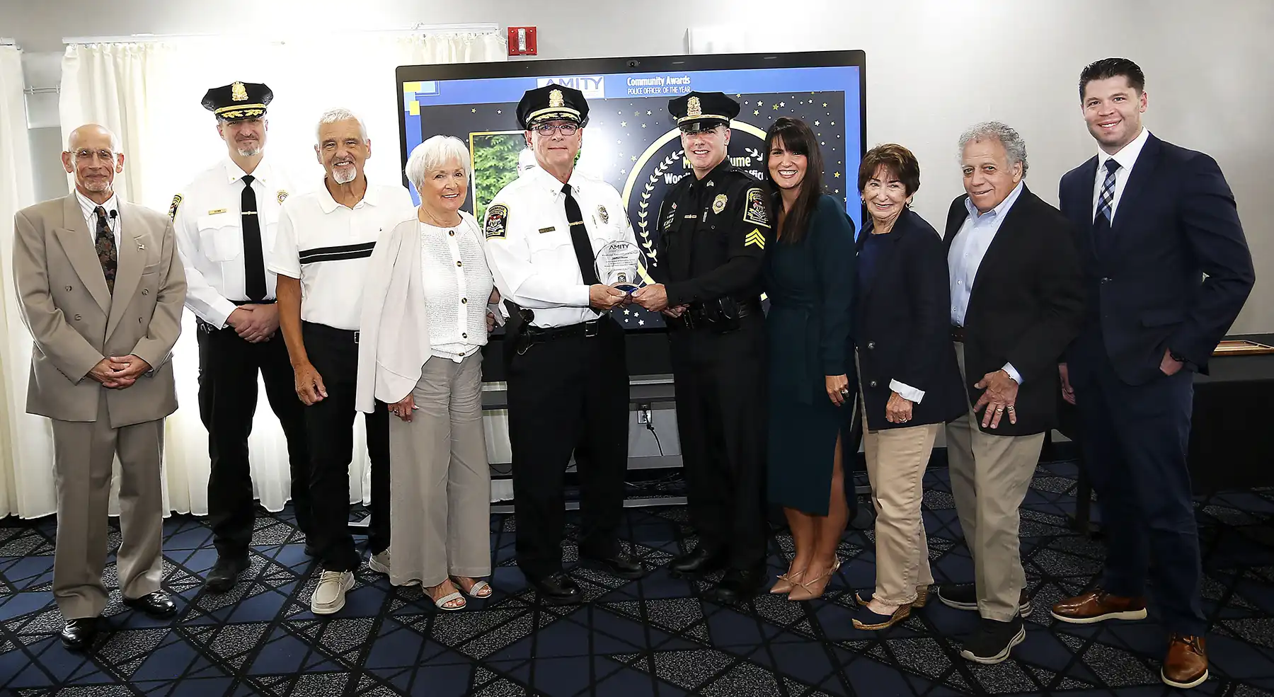 Two police chiefs in white shirts present a crystal award to a uniformed officer, smiling with a group of people at the Amity Chamber event.