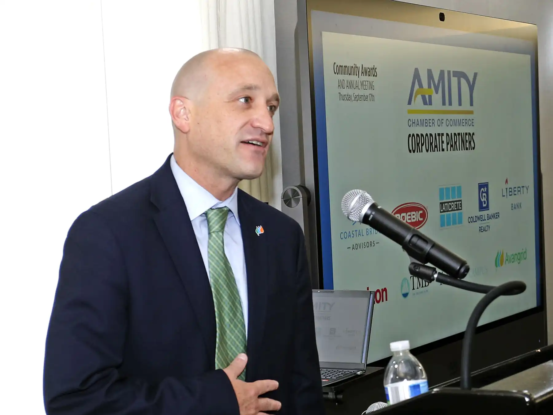 Ted Novicki in a dark suit with a green tie speaks into a microphone at a podium, with an Amity Chamber of Commerce logo on a large screen behind him.