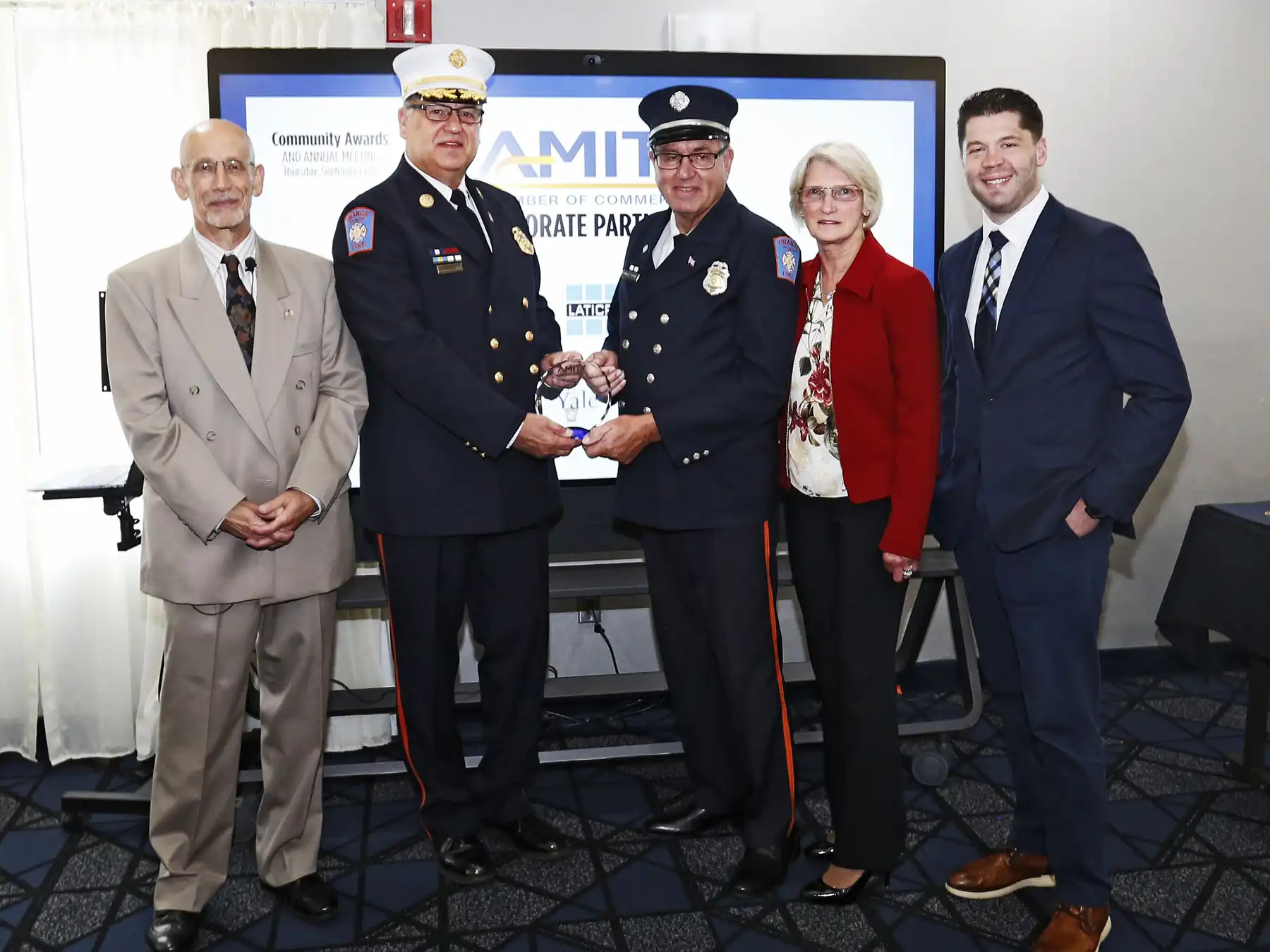 A fire chief in a white hat presents an award to a firefighter in a dark uniform, standing with a man in a beige suit and a woman in a red jacket.