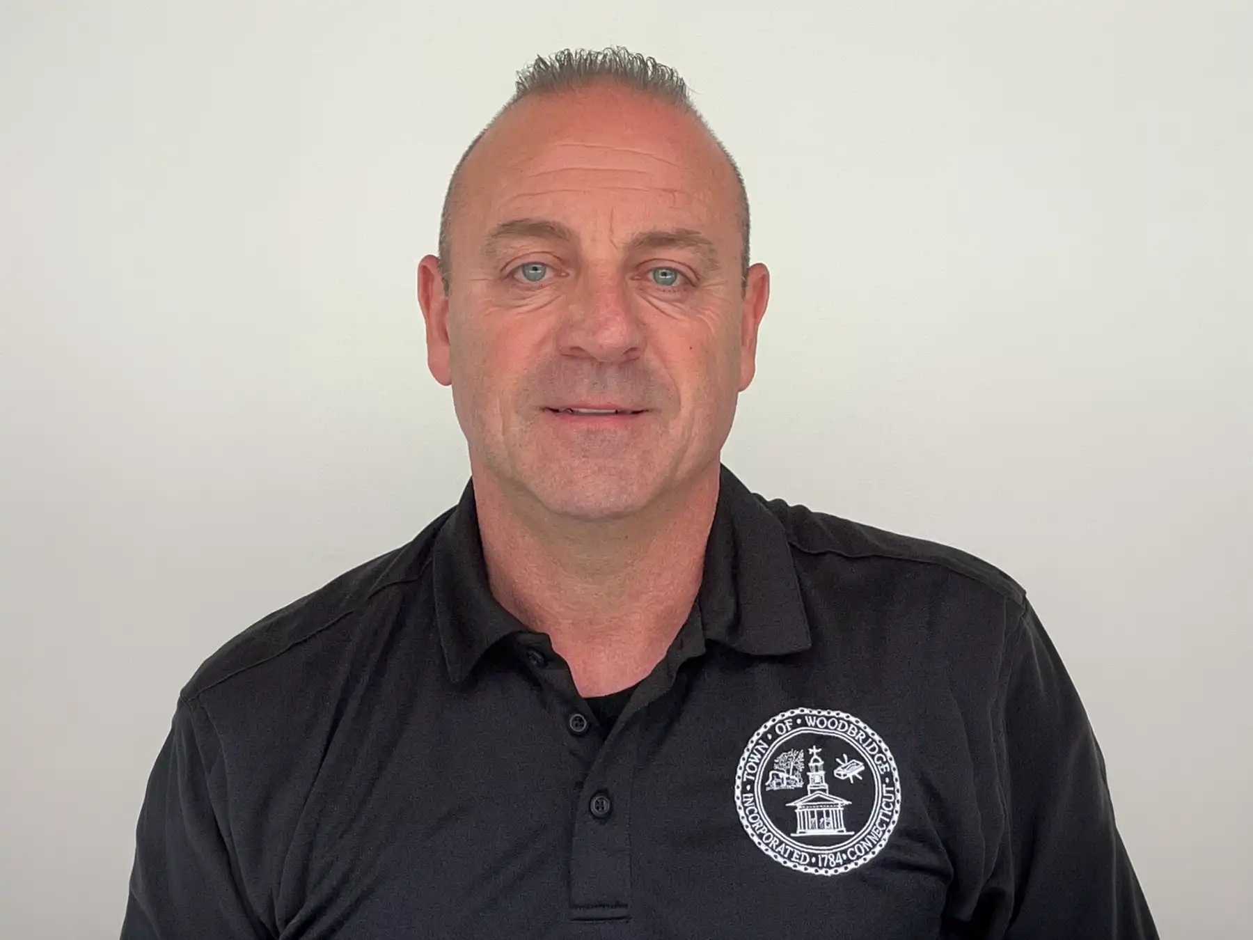 A head and shoulders portrait of a smiling man, Michael Siciliano, wearing a black polo shirt with the official Town of Woodbridge, Connecticut seal.