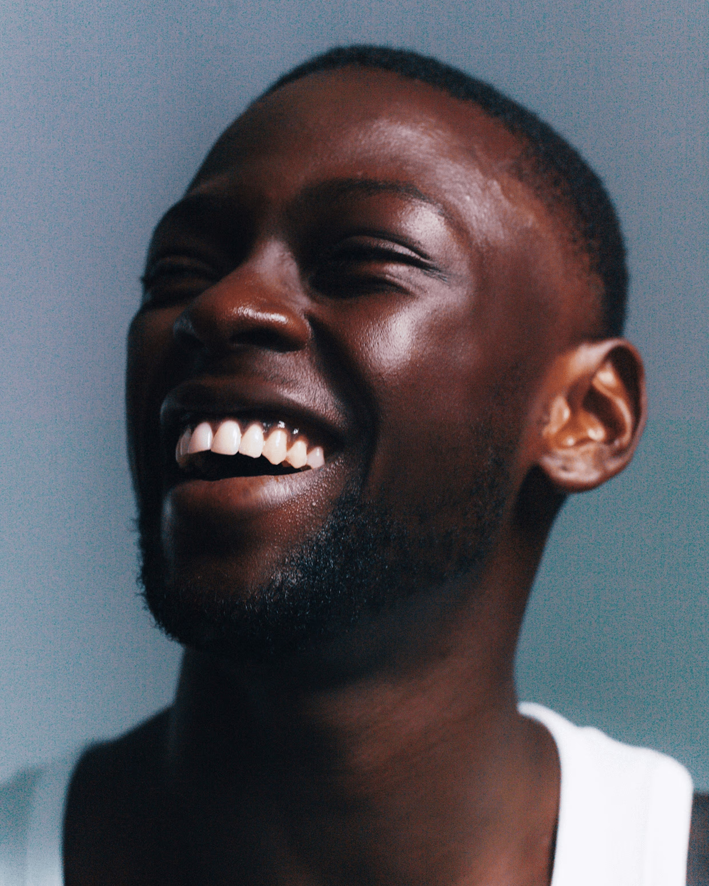 Close-up of a smiling man with short hair and beard wearing a white shirt against a plain background.