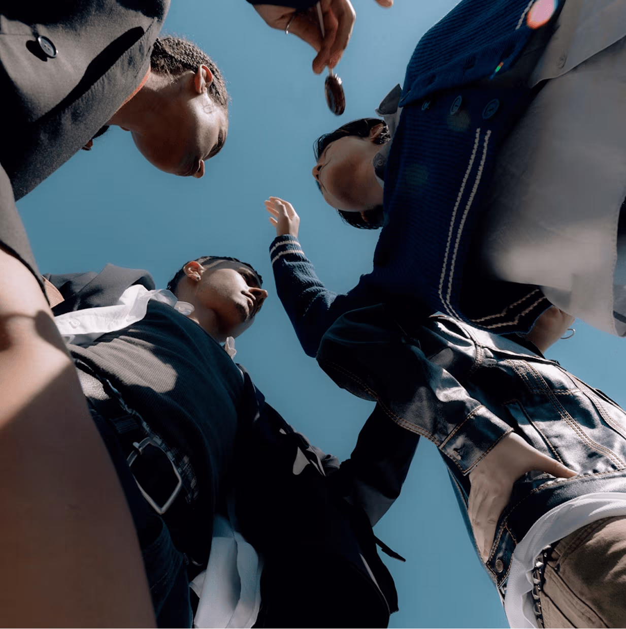 Person wearing colorful rings and an orange sweater typing on a MacBook Air with stickers, beside a croissant, iced drink, vintage Nikon camera, and white earphones on a wooden table.