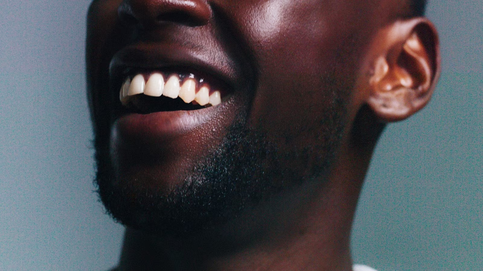 Close-up of a smiling man showing his white teeth and beard on a blue-gray background.