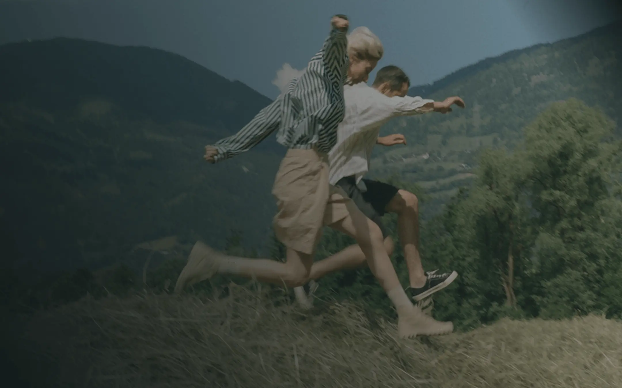 Two people running and jumping outdoors on a grassy hill with mountains and trees in the background.