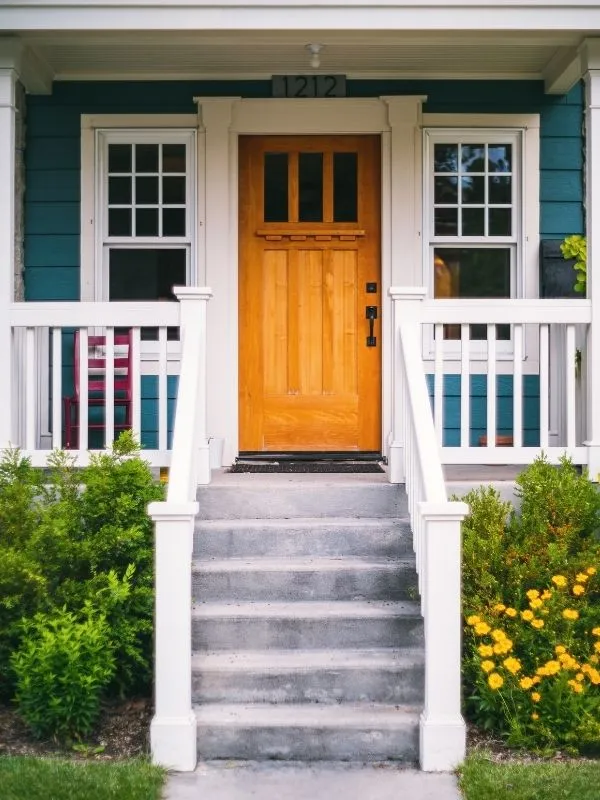 Front porch with concrete steps leading to a wooden door flanked by white railings and green bushes with yellow flowers.