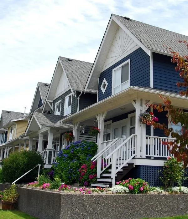 A row of colorful suburban houses with porches, flower beds, and white railings under a cloudy sky.
