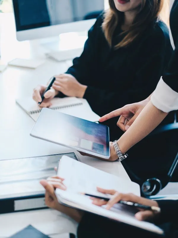 Close-up of two people collaborating over a digital tablet while another person takes notes in a notebook.