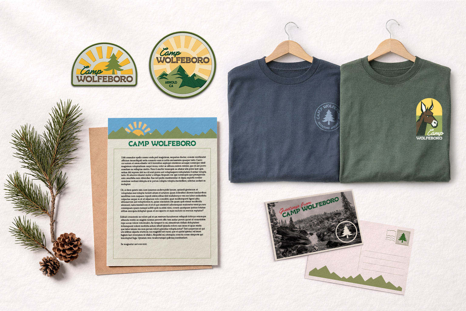 Camp Wolfeboro merchandise including two embroidered patches, two folded t-shirts with camp logos, a letter with envelope, a black and white postcard, and a blank postcard, all placed on a white surface with pine branches and pine cones.