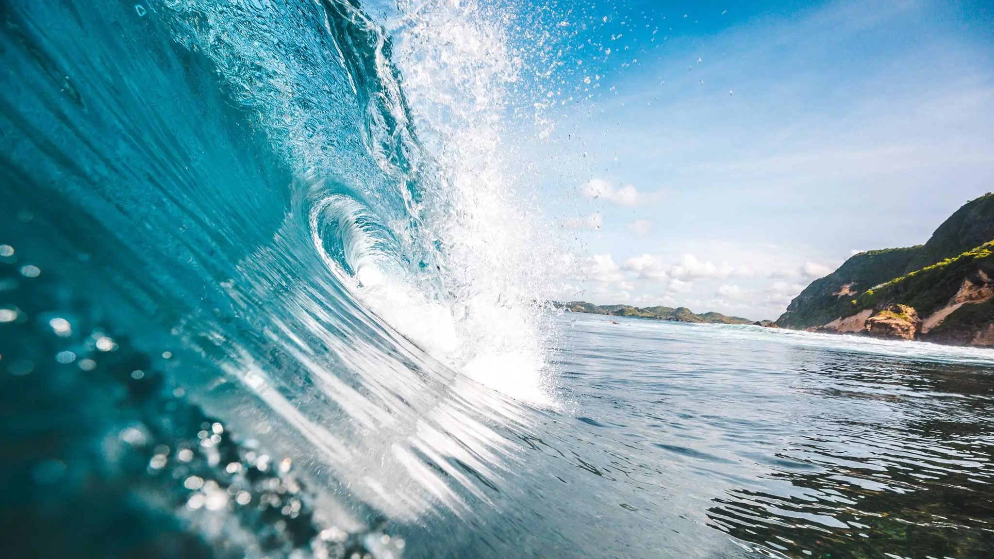 Large turquoise ocean wave barreling with white sea spray and green coastal hills.