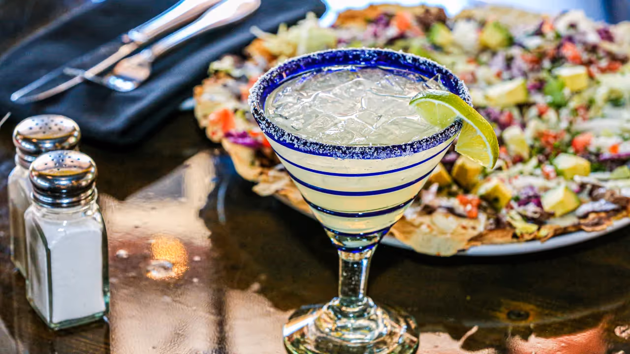 A glass of margarita with a lime wedge garnish in front of a dish of nachos and avocados.