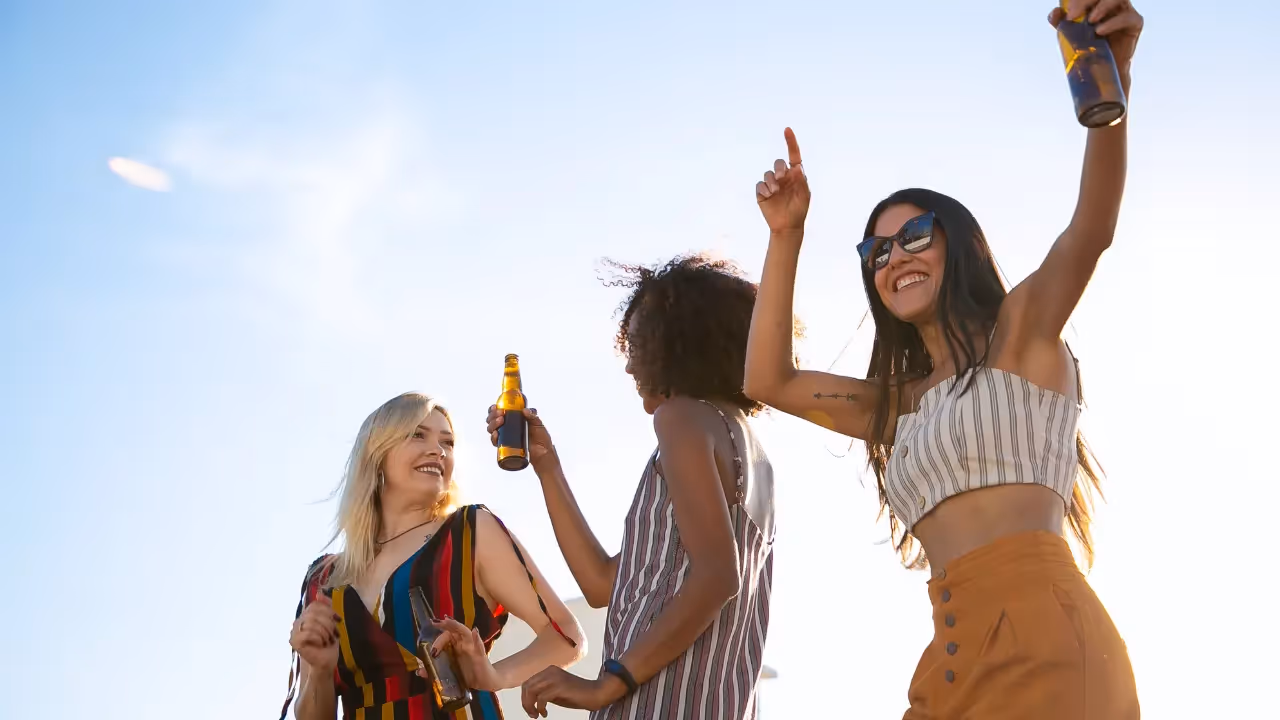 Three women holding glass bottles are dancing against a blue sunny sky.