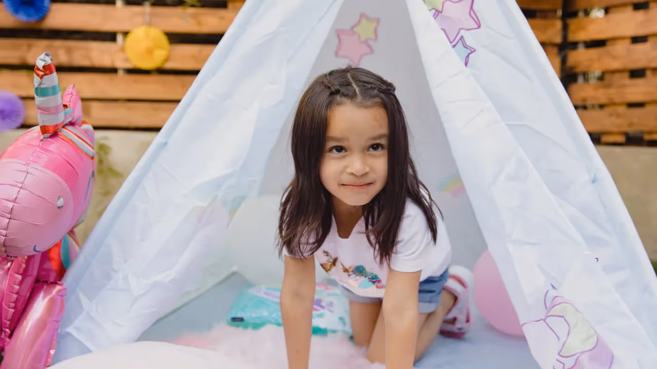 A young girl in a pink tshirt is kneeling in a white canopy tent which is decorated with pink and yellow stars. Next to the tent is a pink unicorn balloon. 