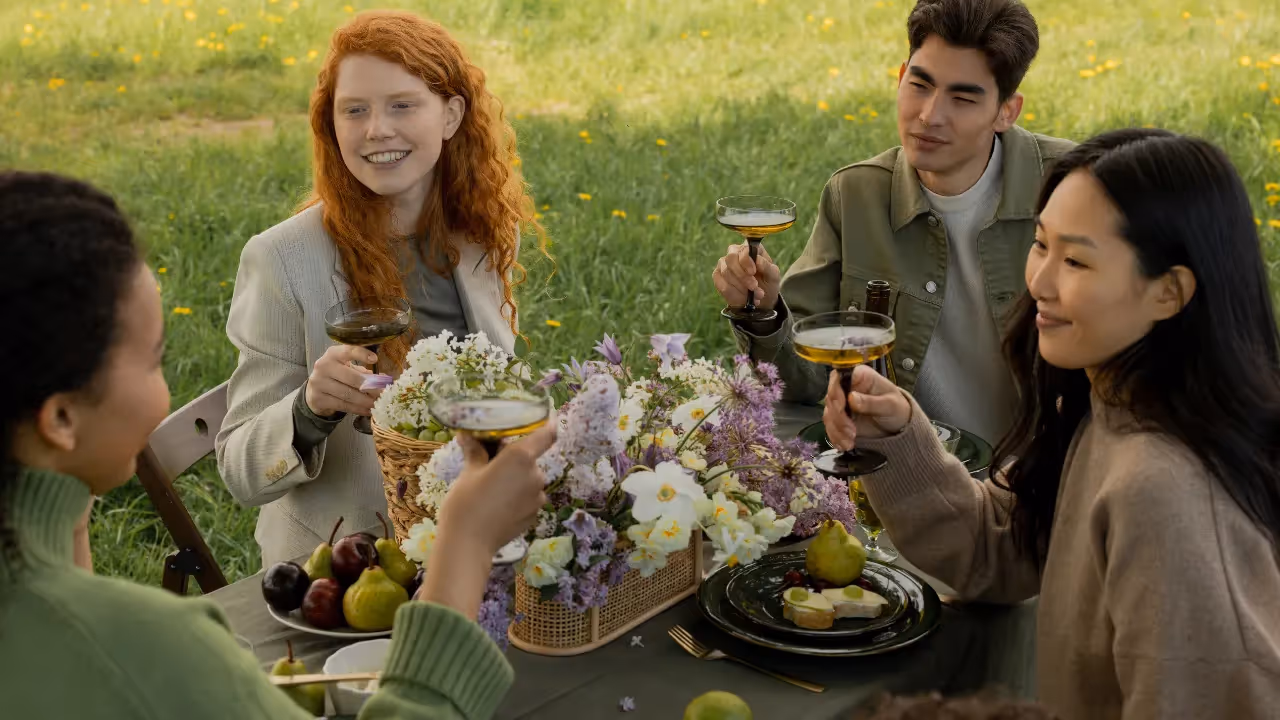 A group of friends sit around a table outside which is filled with flowers and fruits. The group are holding glasses as if to make a toast.