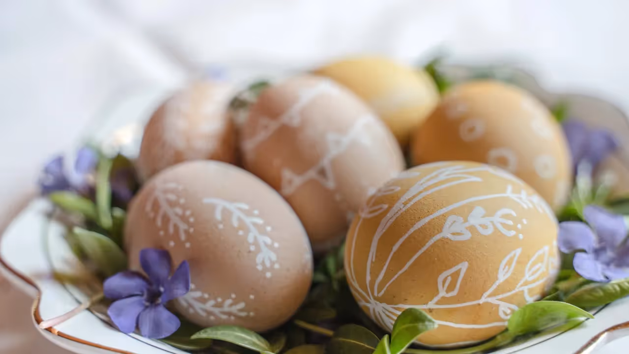 A bowl of painted eggs is surrounded by purple flowers. 
