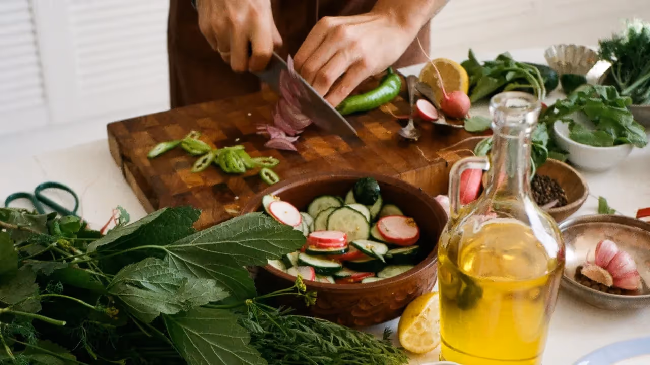Vegetables are being chopped on wooden chopping board. There are lots of bowls of vegetables and a jar of olive oil.