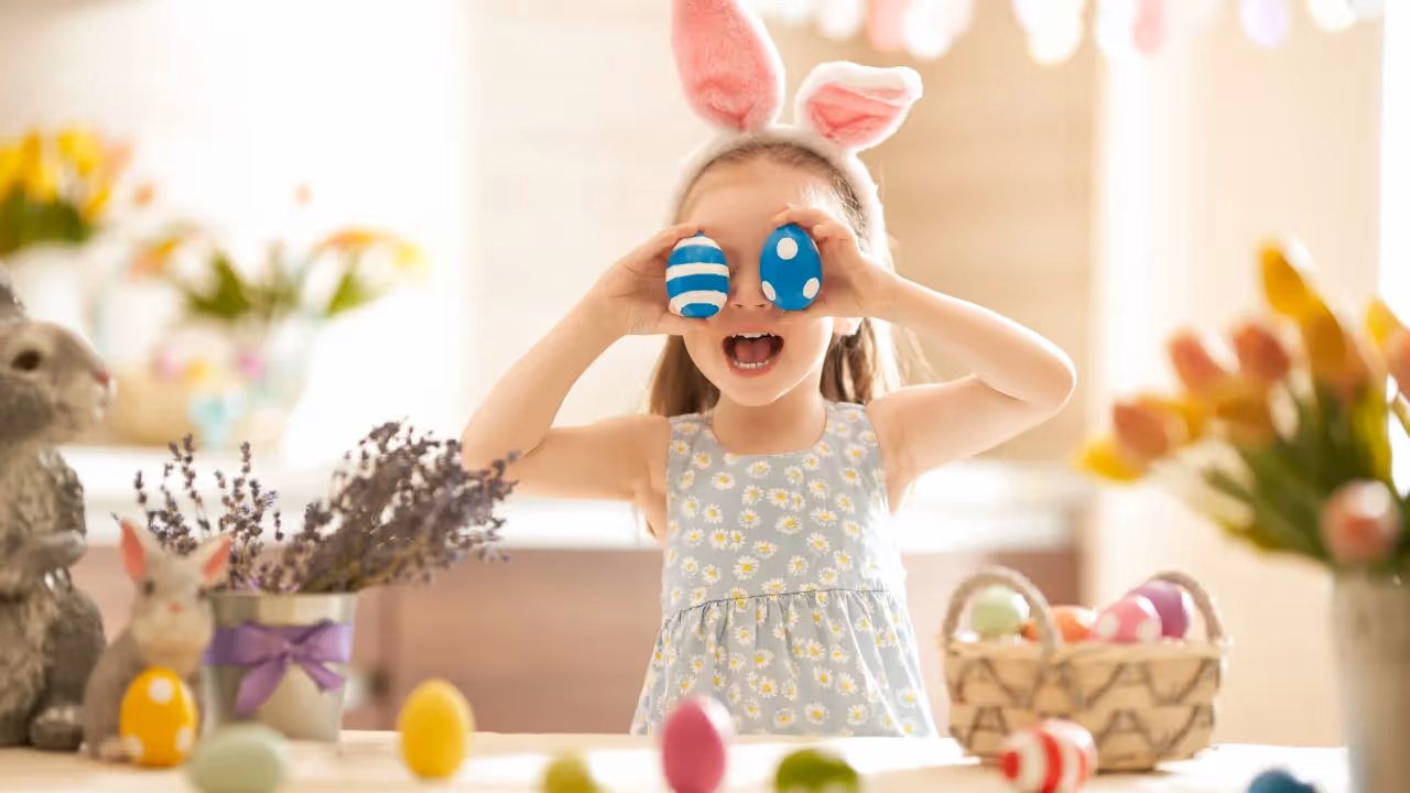 A little girl wearing bunny ears holds two blue easter eggs in front of her eyes. 
