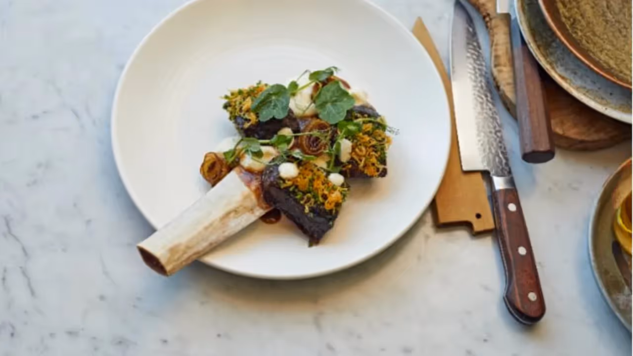 A piece of meat on the bone sits on a white plate next to a sharp kitchen knife. The meat is garnished with green leaves and piped potato.