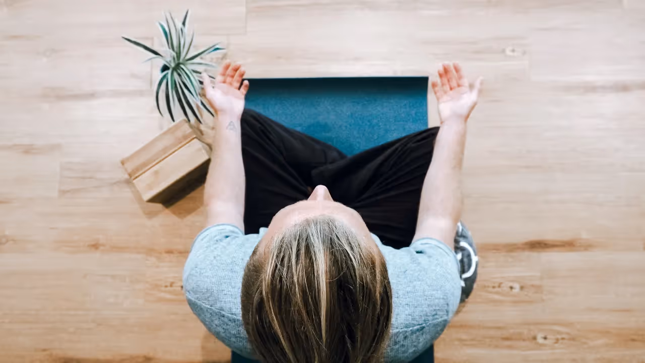 A bird's eye view of a person in a grey top and black trousers sitting on a yoga mat with their legs crossed and their hands balanced on their knees. Next to them is a plant and yoga blocks. 