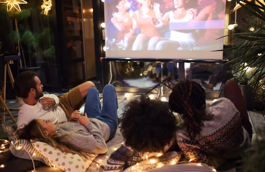 A cosy outdoor movie night setup with friends lounging on cushions under warm string lights, enjoying a private film screening. A unique HOMETAINMENT experience in London, perfect for exclusive home gatherings and personalised entertainment events.
