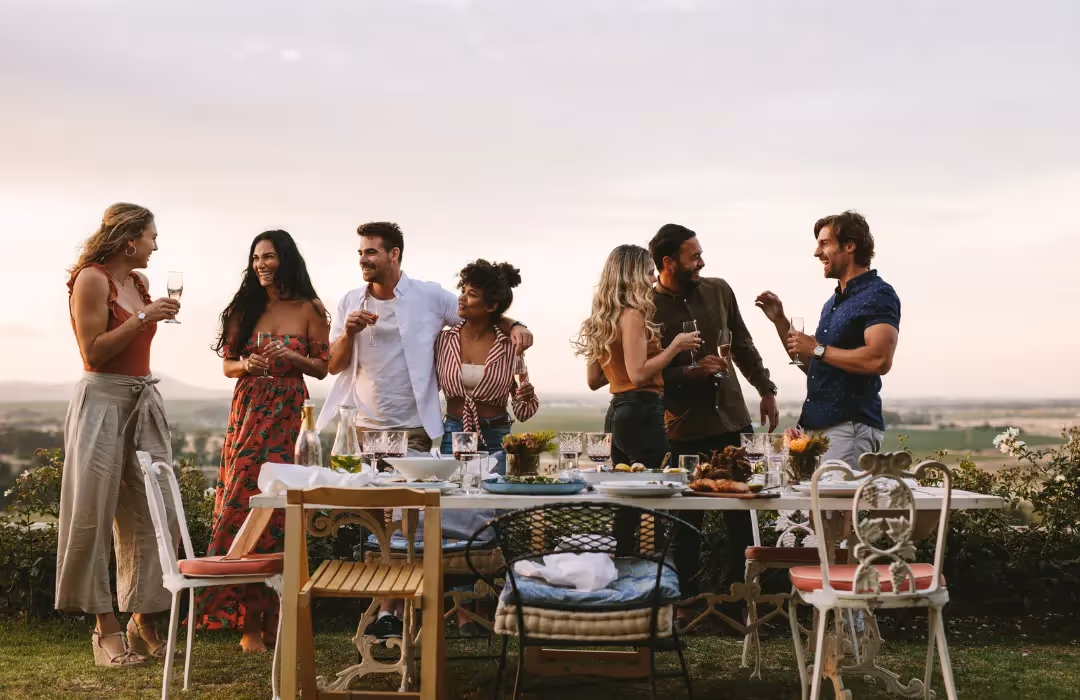 A group of friends enjoying an alfresco dining experience with scenic countryside views, sharing drinks and laughter around a beautifully set outdoor table. A perfect setting for a private home gathering or exclusive event.