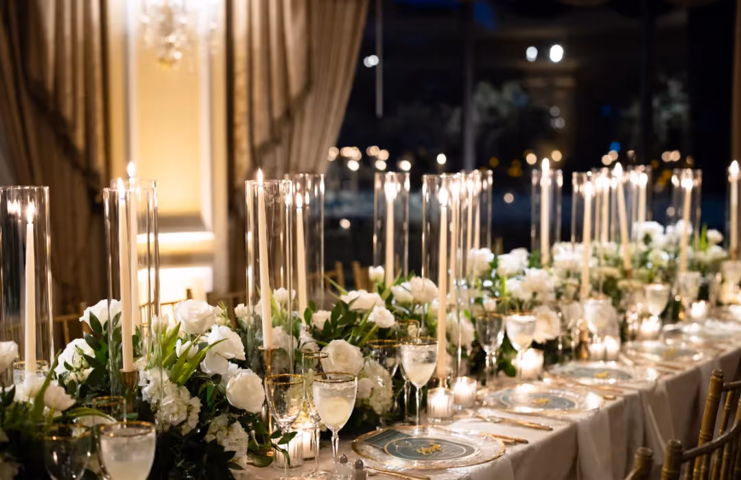 Elegant luxury dining setup featuring a beautifully decorated long table adorned with tall glass-encased candles, lush white floral arrangements, and gold-accented tableware. A perfect ambiance for an upscale private event or celebration at home.