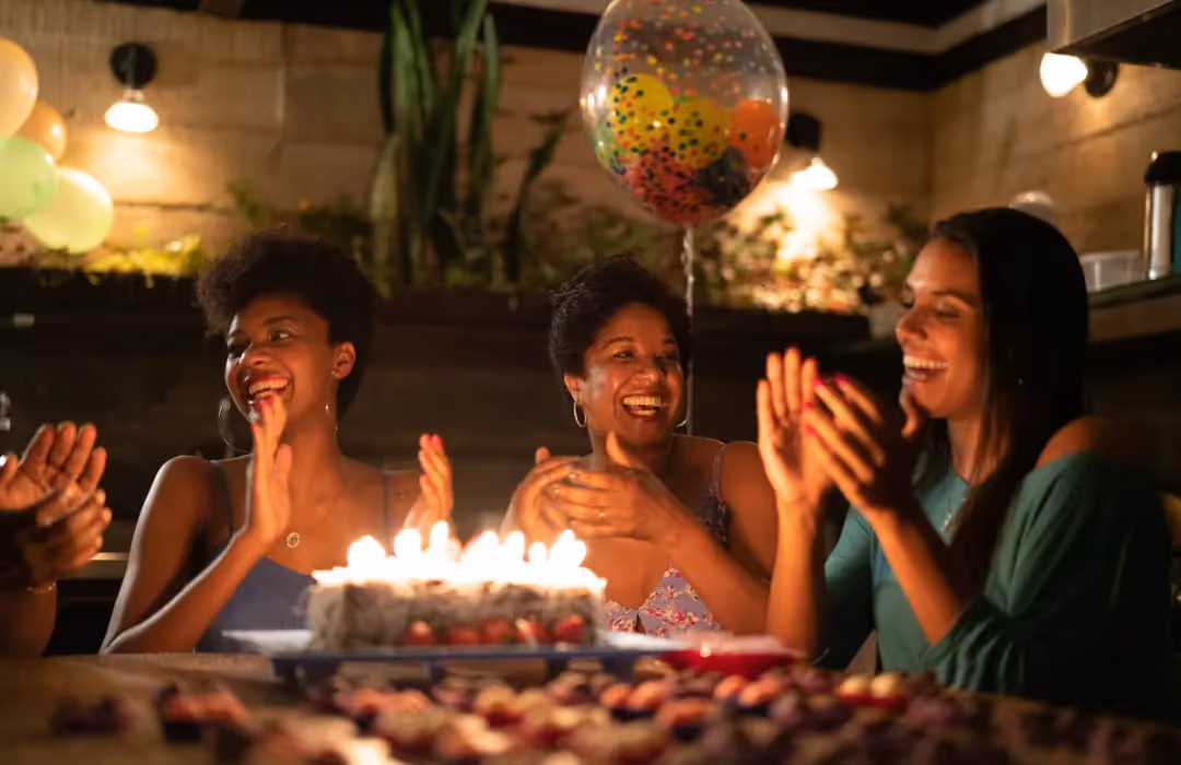 A joyful birthday celebration with friends and family gathered around a candlelit cake, clapping and laughing in a warm, intimate setting. A perfect example of a personalised home event filled with happiness and togetherness.