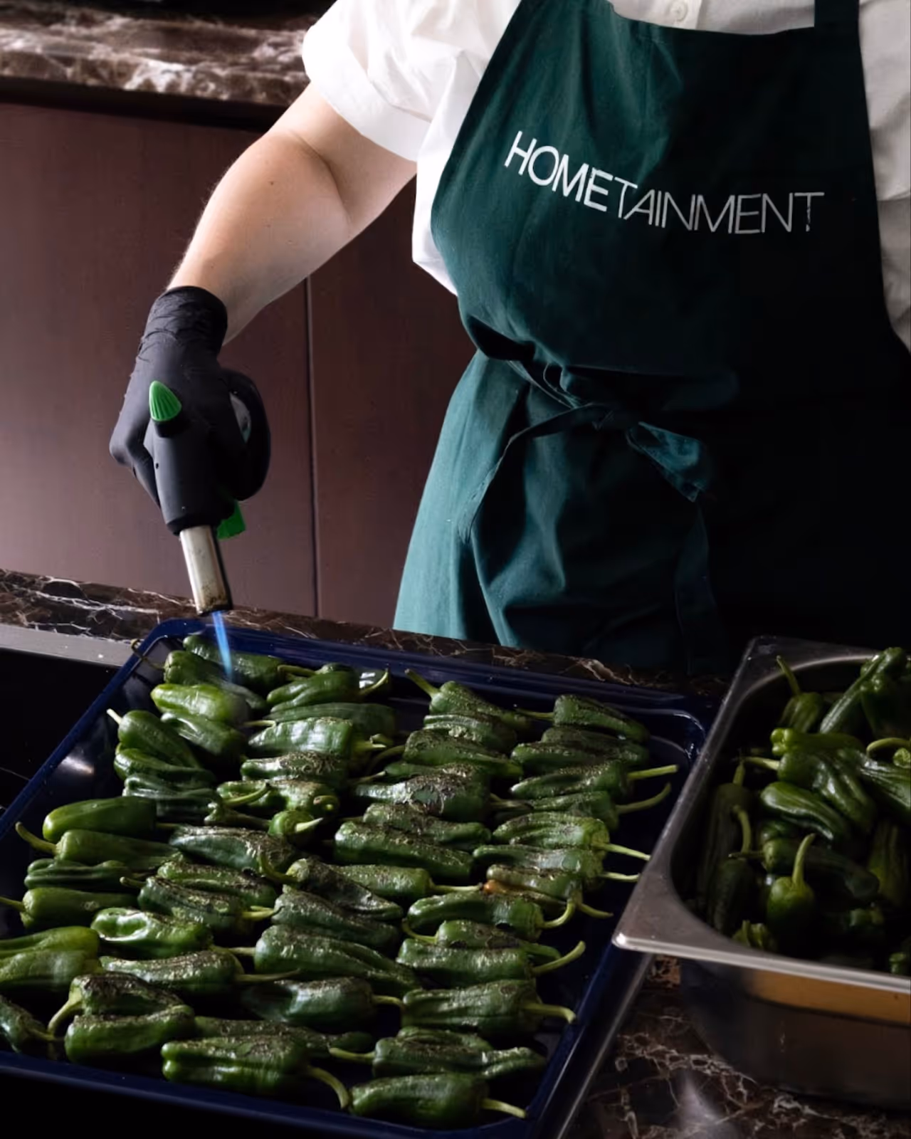 Private chef torching padron peppers in a modern kitchen while wearing a HOMETAINMENT apron during a luxury at-home dining experience in London
