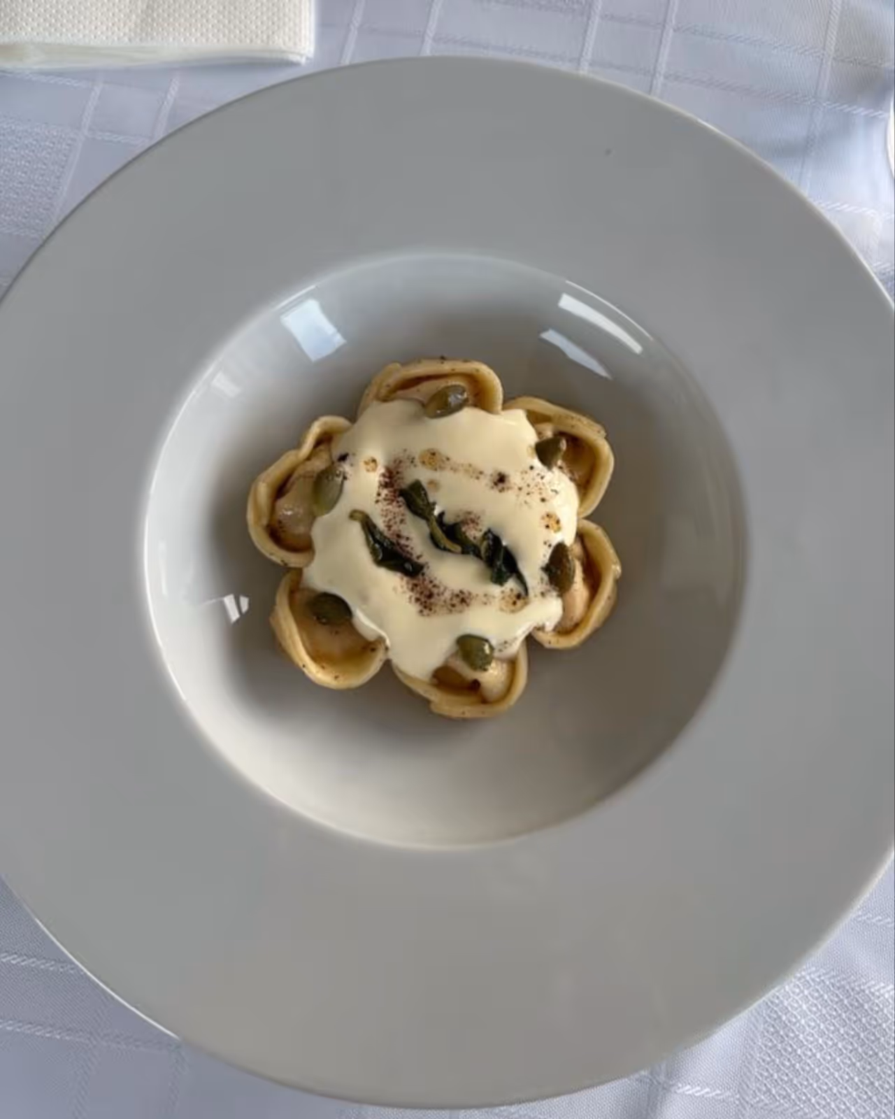 A gourmet dish of flower-shaped tortellini arranged in a circular pattern, topped with a creamy white sauce, capers, and crispy sage leaves. Served on a large white plate over a white tablecloth, the presentation is elegant and minimalist, emphasizing the delicate structure and fine ingredients of the pasta.