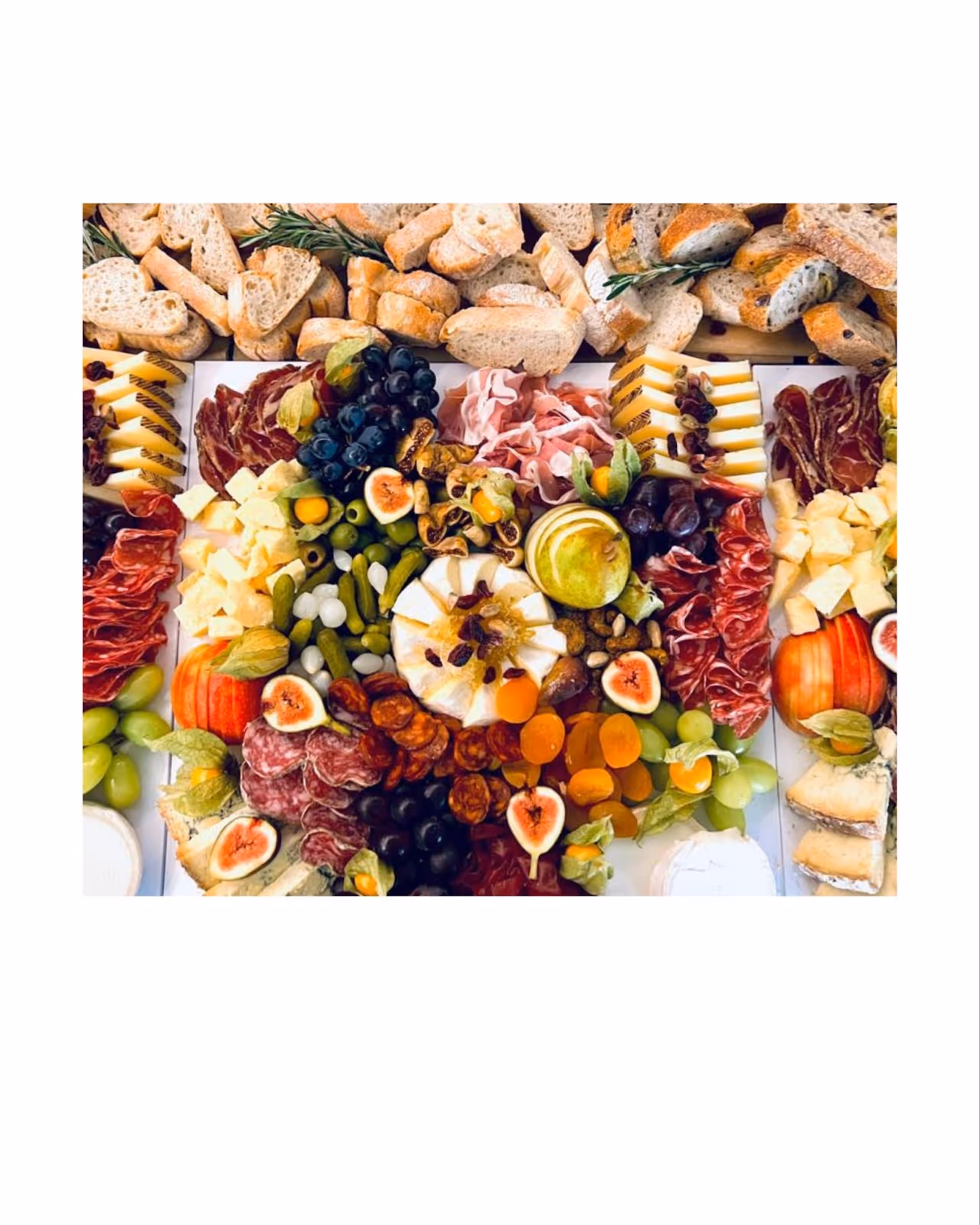 A vibrant charcuterie board filled with an assortment of gourmet cheeses, cured meats, fresh and dried fruits, olives, nuts, and sliced bread. The centerpiece features a round cheese topped with dried cranberries, surrounded by figs, grapes, pears, apricots, and salami, with baguette slices arranged neatly at the top. The display is colorful, abundant, and artfully arranged for an elegant presentation.