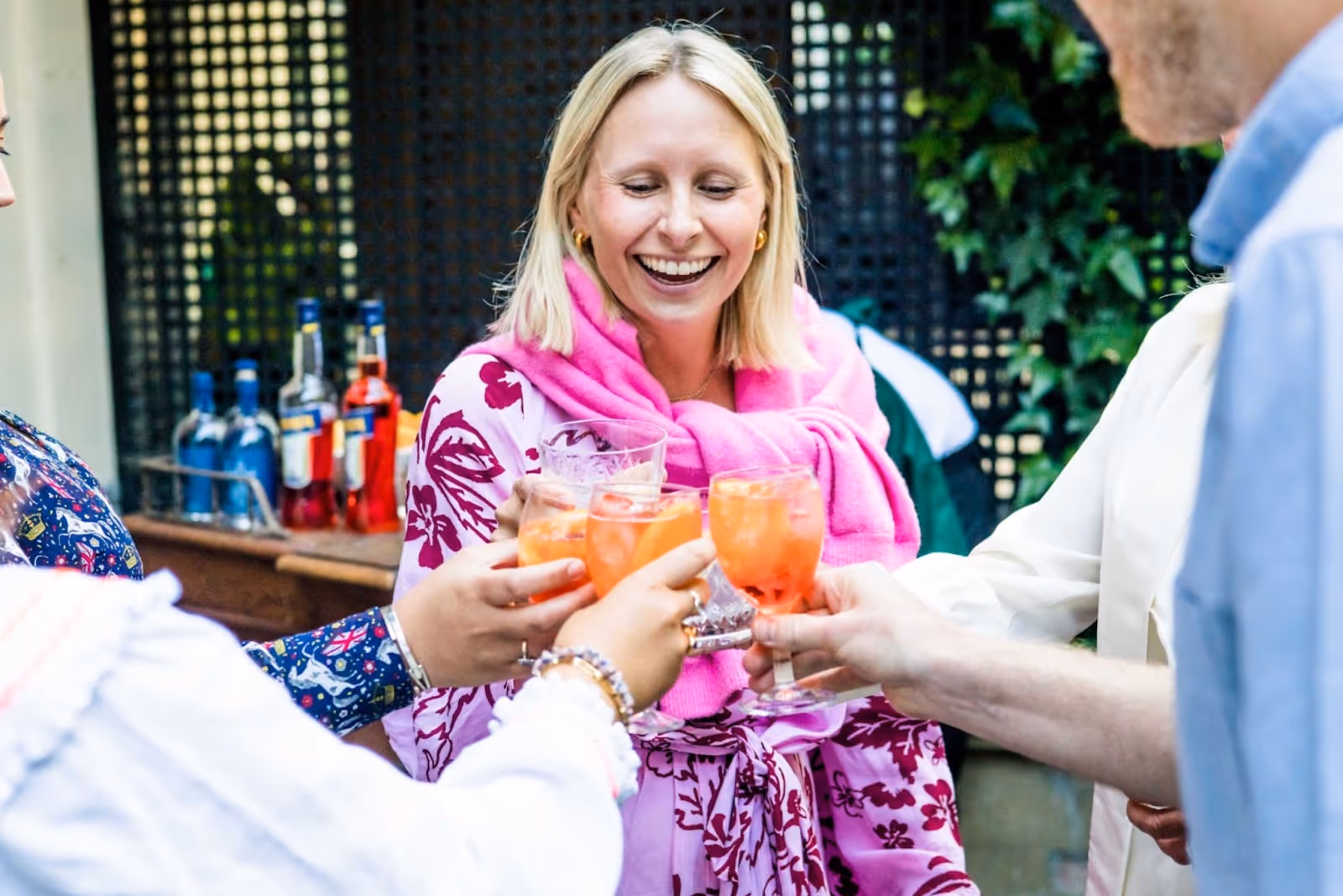 A blond woman dressed in pink in a garden in London cheersing her Aperol Spritz with others at a garden party.x