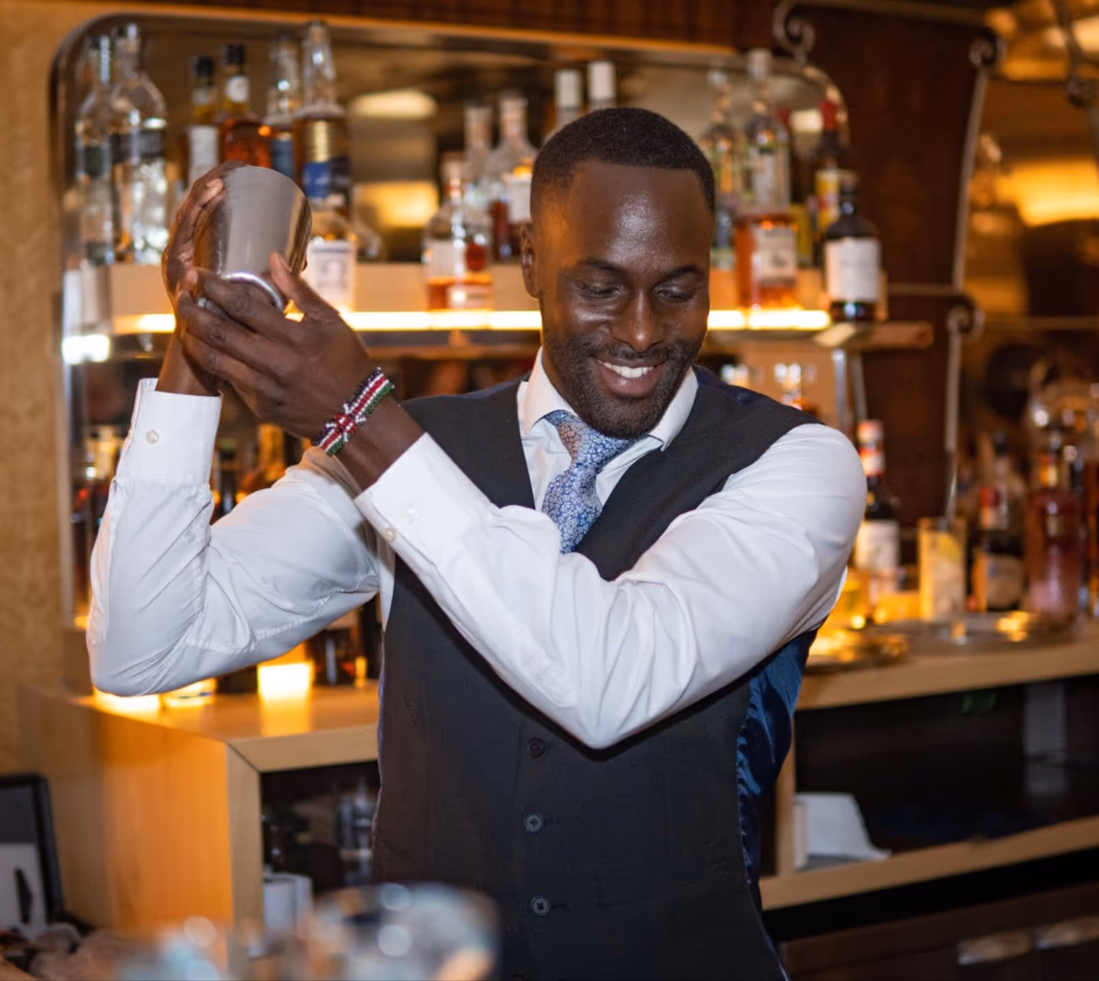 A smiling bartender in a waistcoat and tie confidently shakes a cocktail shaker behind a bar stocked with bottles, creating a lively and professional atmosphere.