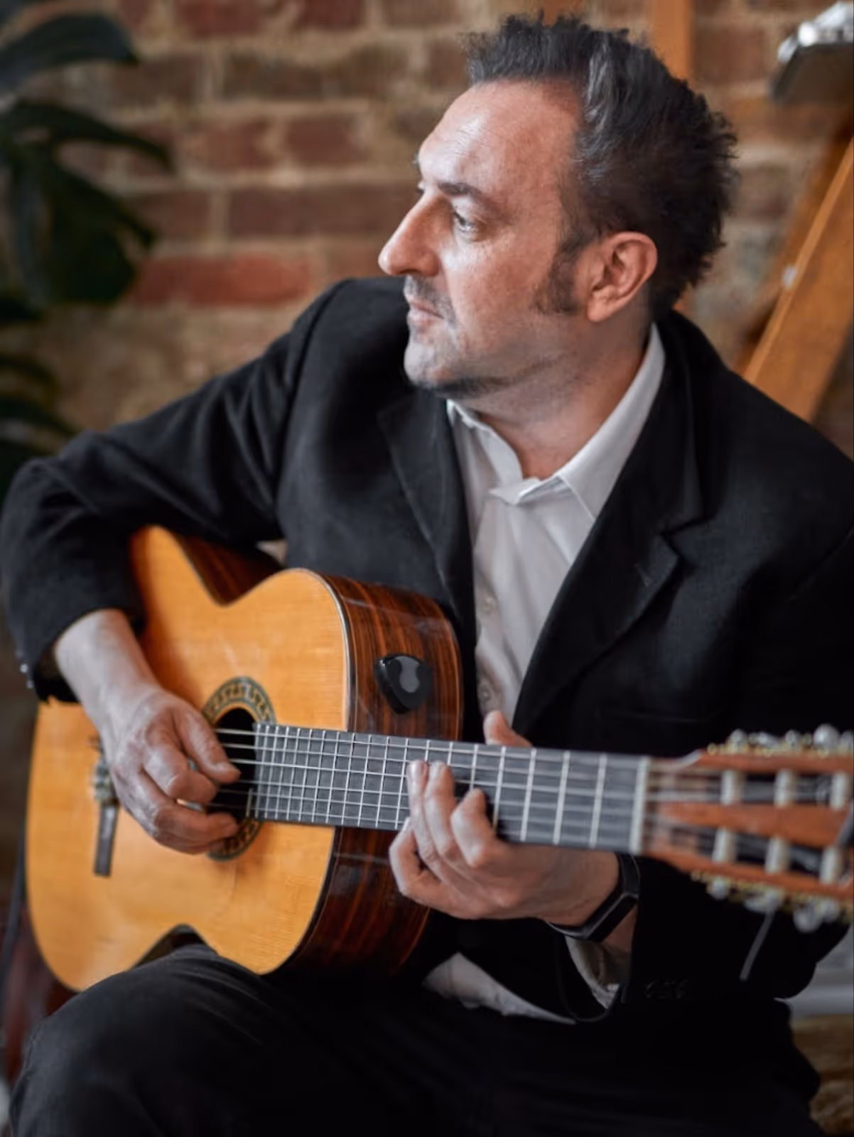 Spanish man playing Spanish guitar with dark hair in a black jacket inside.