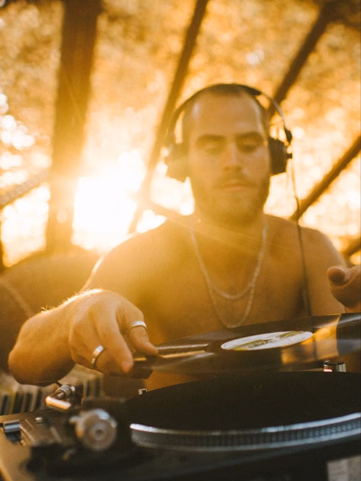 A young DJ putting on a new disk on the decks with headphones and shirtless at a outdoor festival with lots of sun