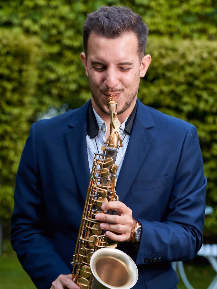 A man smiling whilst playing the sax in a garden in a blue suit jacket.