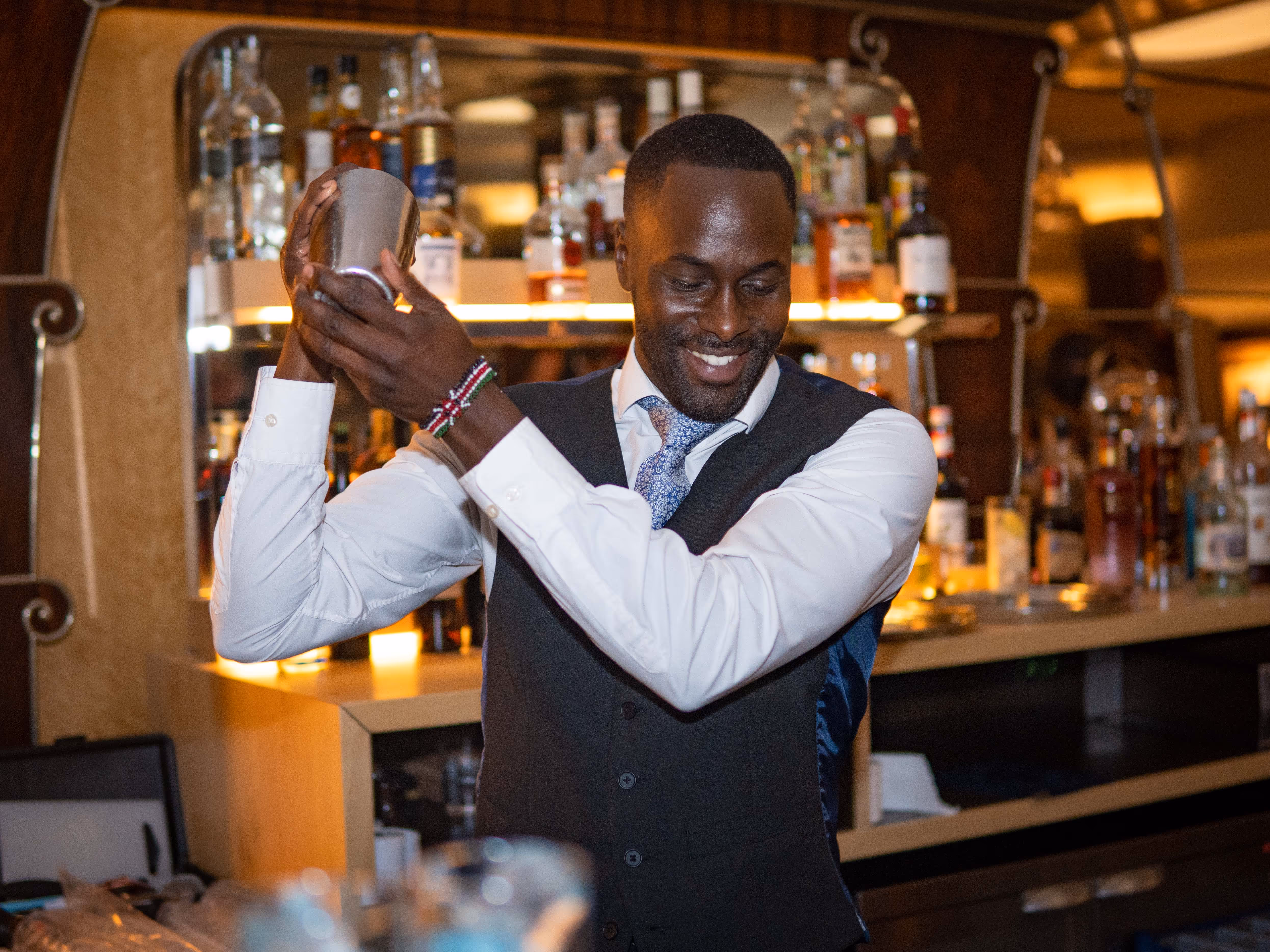 A smiling bartender in a white shirt and dark waistcoat is shaking a cocktail shaker behind a well-stocked indoor bar. The setting is warmly lit, and he is wearing a bracelet with red, green, and black beads.