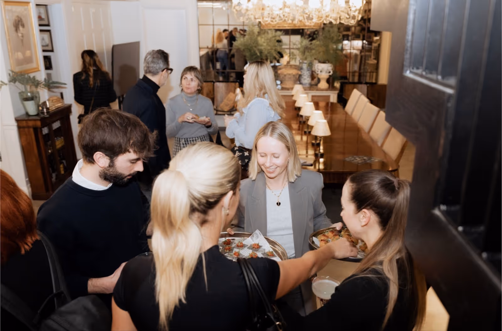 A Blond woman opening a door with platters of food in her hands serving three guests dressed in black in a chic home.