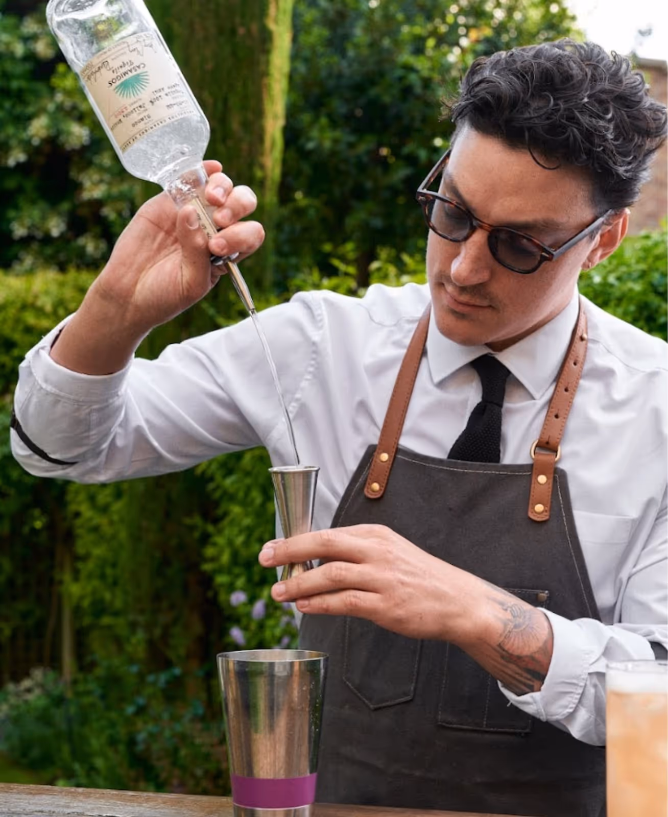 Bartender in a white shirt and black tie with curly hair and round sunglasses pouring some clear alcohol into a silver jigger in a green garden. 