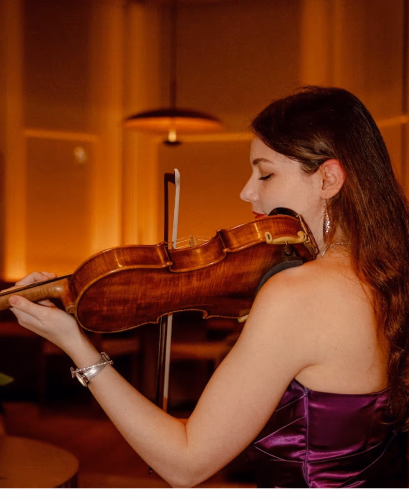 A woman in a strapless purple dress plays the violin in a warmly lit indoor setting, eyes closed as she focuses on the music.
