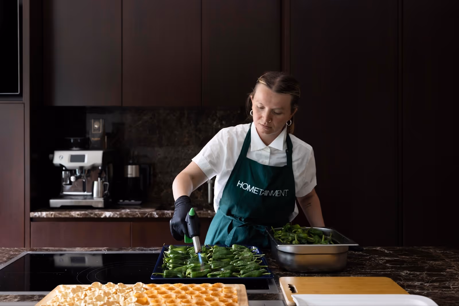 A private chef wearing a HOMETAINMENT apron uses a blowtorch to char green peppers in a modern kitchen, with trays of ingredients on the counter.
