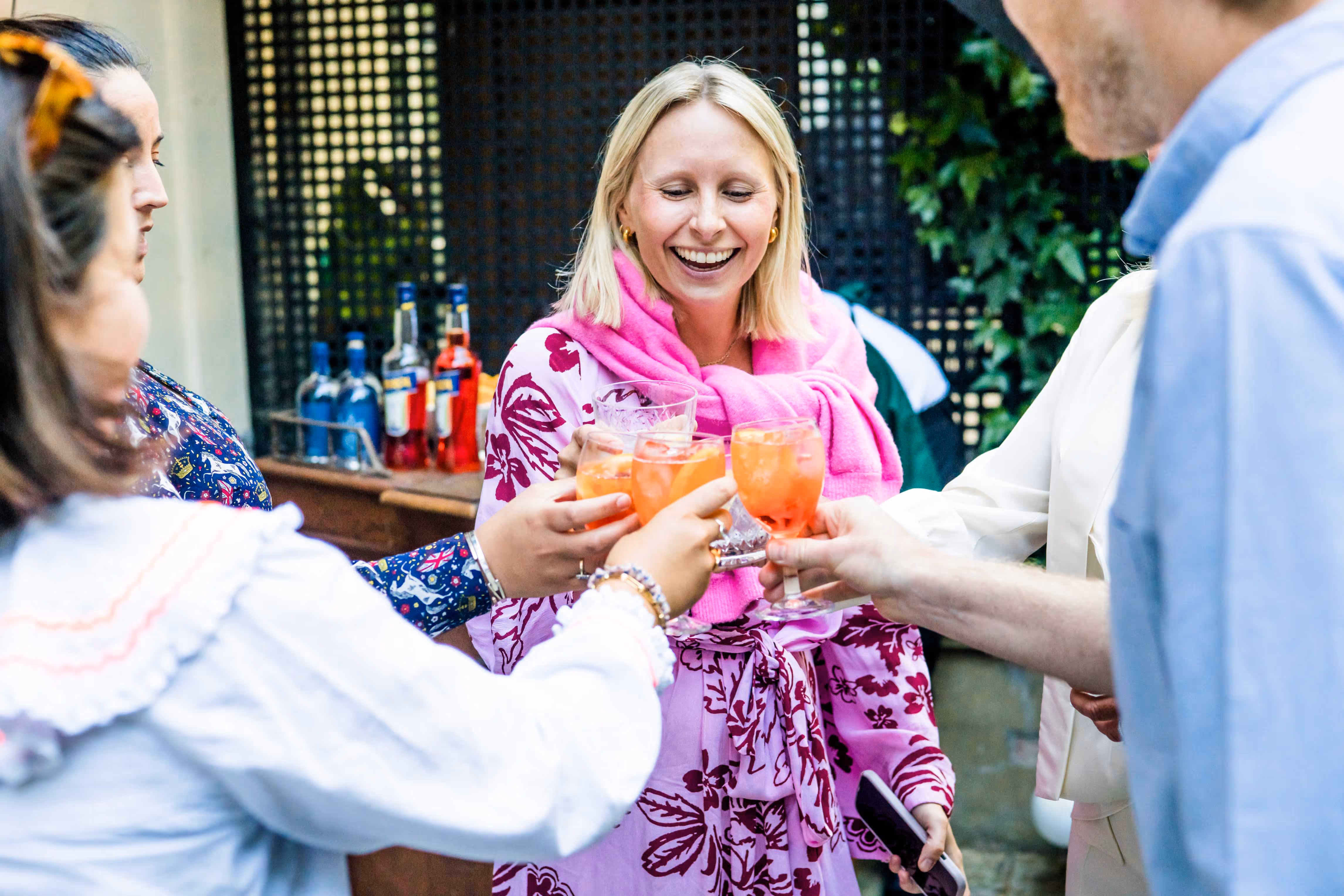 A group of people cheerfully clinking glasses with orange cocktails at an outdoor gathering. A woman in the center, smiling, wears a pink floral dress with a bright pink sweater draped over her shoulders.