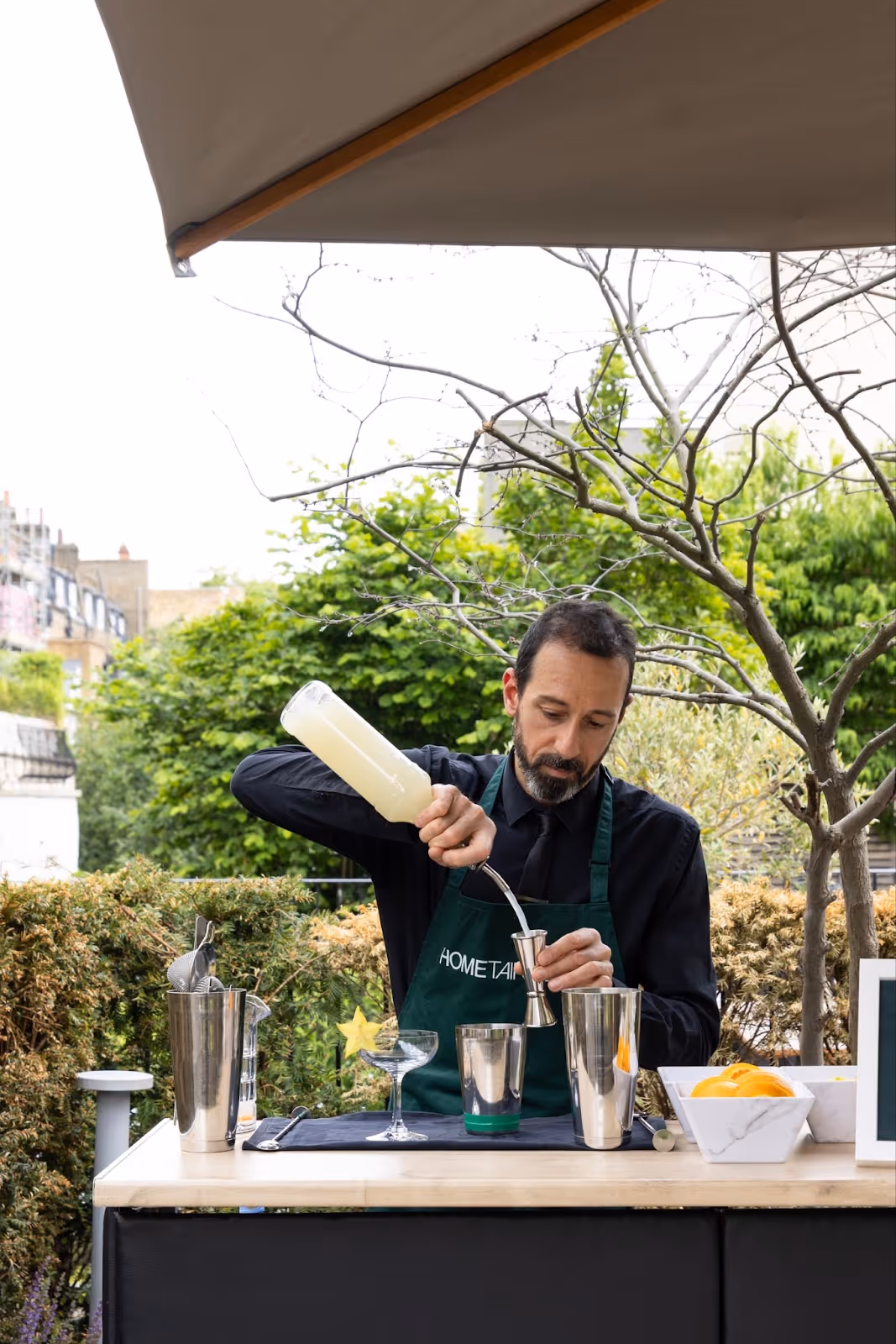 A bartender in a Hometainment apron pouring alcohol into a silver jigger under an umbrella, in a green garden
