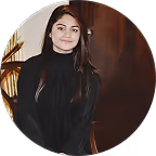 Young woman with long dark hair wearing a black turtleneck sweater seated indoors with a wooden backdrop and a plant.