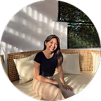 Woman with long dark hair smiling, sitting on a wicker bench with cream cushions in sunlight.