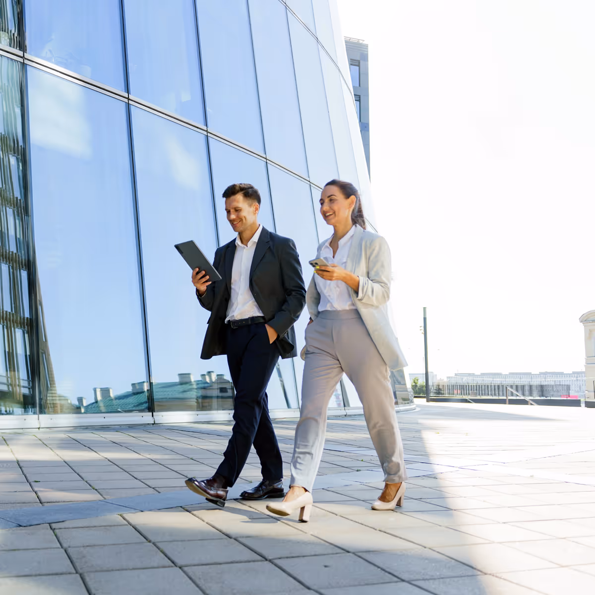 Two business professionals walking outside a modern glass building, looking at digital devices and smiling.