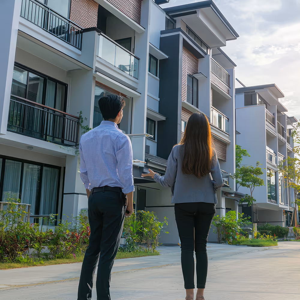 A man and a woman stand on a residential street looking at modern multi-story townhouses with balconies and greenery.
