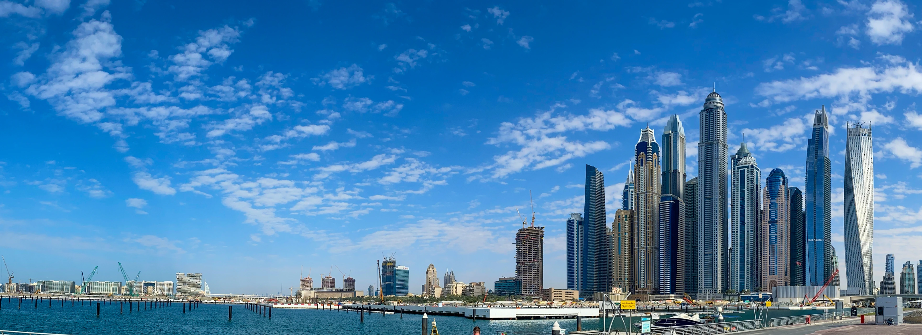 Panoramic view of Dubai city center skyline with tall skyscrapers under a partly cloudy blue sky and calm waterfront.